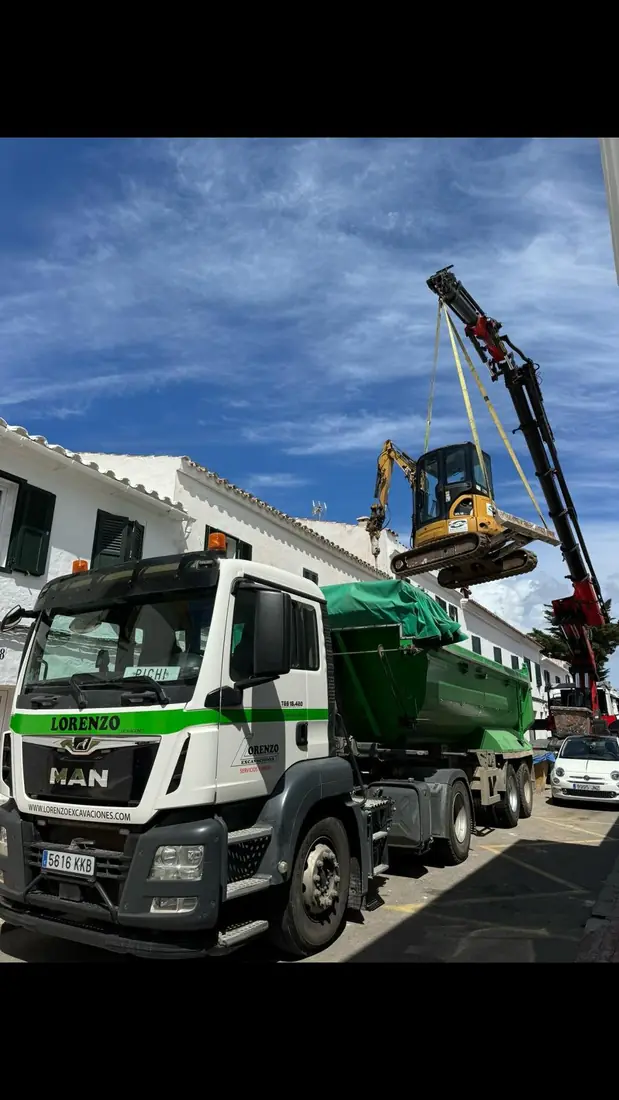 Camión grúa elevando una miniexcavadora por encima de una vivienda